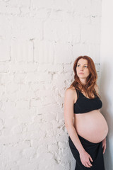 Pregnant woman with red hair in black clothes on a white wall
