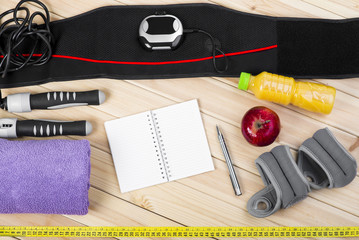 Electrical Muscle Stimulation Belt, Jump Rope, Ankle Weights, Towel, Tape Measure, Apple, Bottle Of Juice, Notepad To Workout Or Diet Plan On Wooden Floor. Sport Fitness Background.
