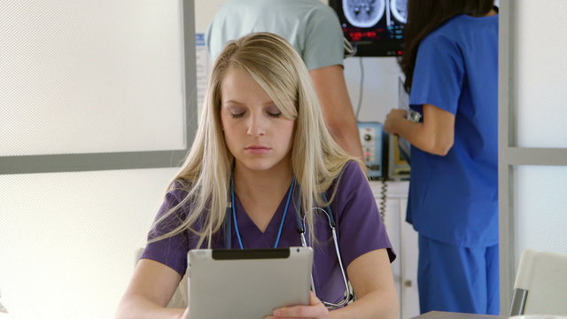 Pretty Blond Nurse With Other Medical Staff Members Reviewing Data On Monitor In Background Looks Up From Her Tablet Pc And Smiles. Scene Dollies Left