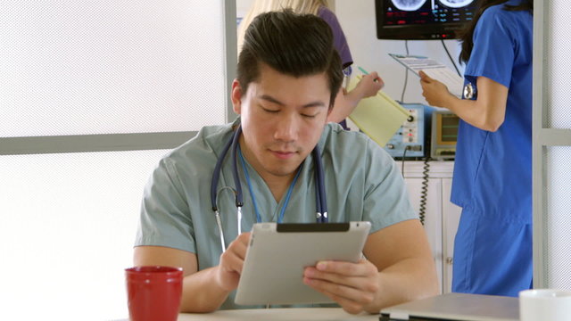 Young Asian doctor or medical intern sitting in hospital lounge stops using his table and smiles while other staff members work in background.
