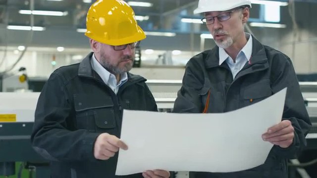 Two engineers in hardhats discuss a blueprint while standing in a factory. Shot on RED Cinema Camera.