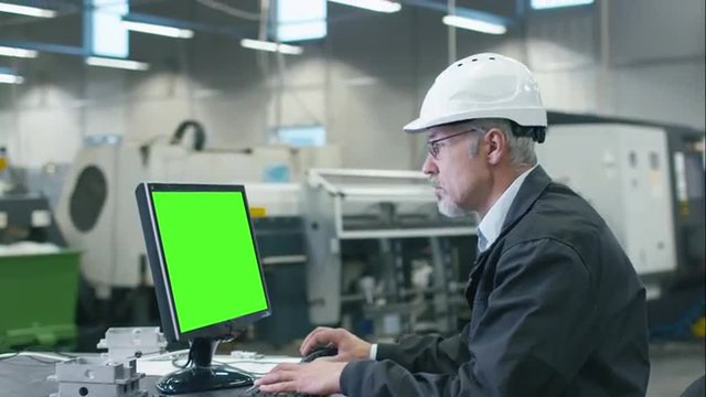 Senior engineer in glasses is working on a desktop computer with a green screen on monitor in a factory. Shot on RED Cinema Camera.