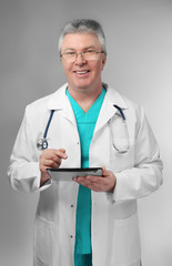 A handsome doctor with stethoscope and tablet standing on grey background