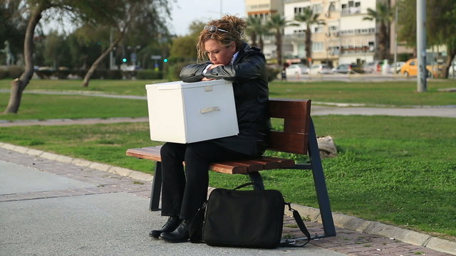 A fired businesswoman in a suit with a box of personal items sitting on the park bench
