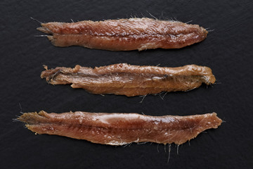 Three anchovy fillets with oil arranged symmetrically and isolated on black textured wood from...