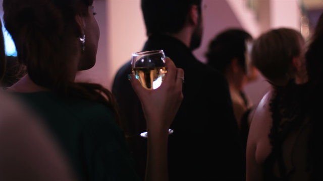 Young attractive female standing back to the camera and keeping a glass of white wine at the party and watching at the stage where a performance is going on. 