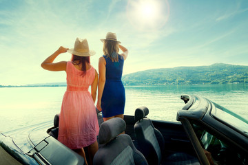 Zwei Frauen stehen im offenen Cabrio und schauen auf den See bei Sonnenuntergang