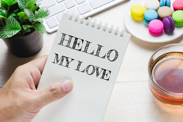 Man holding HELLO MY LOVE message on book and keyboard with a hot cup of tea, macaroon on the...