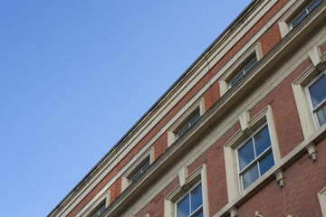 The facade of an old, red brick building with blue sky above. The colonnade of a period building with rows of uniform windows. A view from the ground looking upwards.