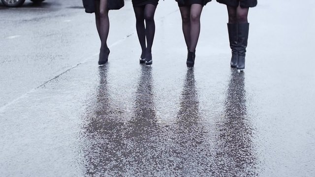 Legs of four woman in shoe on heels walk in lockstep on wet asphalt. Slow motion