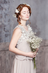 Young red-haired bride in pearl-gray dress. She stands, her eyes are dreamy closed, she is holding a bouquet of white wildflowers. White flowers in her hair.