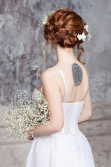 Young red-haired bride in elegant wedding dress. She stands with her back to the viewer. She is holding a bouquet of white wildflowers. White flowers in her hair. She has a mandala tattoo on her back.