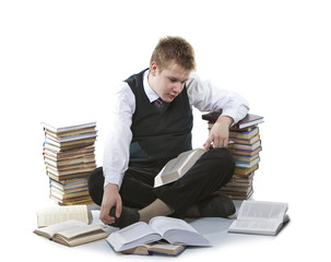 The schoolboy in a school uniform sits on a floor, near to packs of books, with the opened book in hands