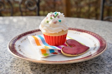 Cupcake or cup cake with whipped cream and small colorfull sugar perils served on the small dessert plate with paper napkin and red heart. Sweet is on the granit garden table outside.