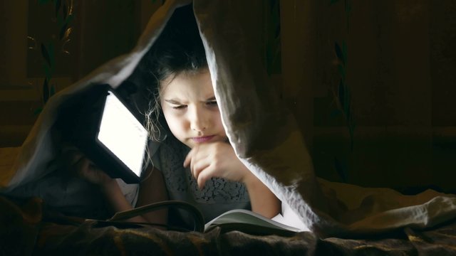 girl  reading book under covers with flashlight at night