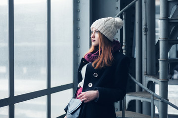 Portrait of a young beautiful elegant redhead woman wearing stylish winter clothes. Model looking through the window. Female fashion concept. Closeup