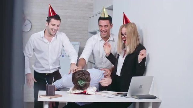 Office workers in party hats smashing face of their colleague into the birthday cake, clapping hands and laughing