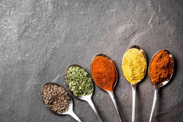 Colorful spices on metal spoons on black rough rock surface.