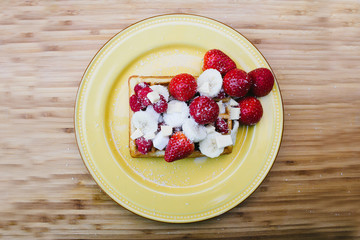 Cenital view of a waffle with strawberries, raspberries, sliced banana and powdered sugar