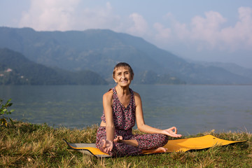Old Happy Woman 68 years old smiling and doing yoga exercise in morning near mountain lake