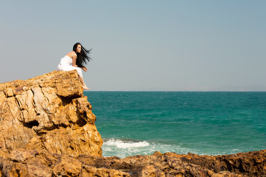 Young Woman sitting outdoor on rock beach, looking on blue ocean, enjoy freedom