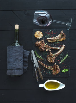 Grilled lamb chops. Rack of Lamb with garlic, rosemary, spices on slate tray, wine glass, oil in a saucer and bottle over black wood background