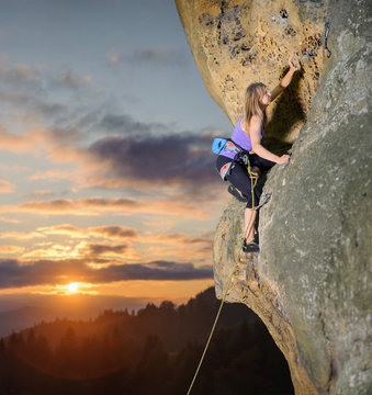 Young female rock climber on a cliff face against scenic sunset background. Reaching for the next carbine.