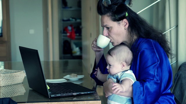 Young, tired mother in bathrobe trying work on laptop with her little child 
