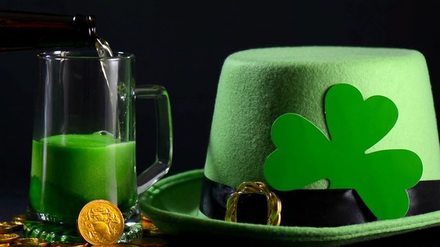 St Patricks Day pouring green beer with green leprechaun hat, gold coins and shamrock against black background, zoom.