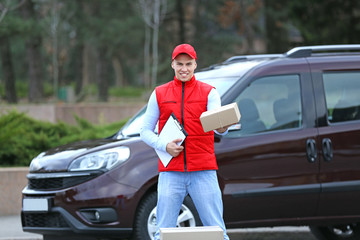 Young handsome delivery man with box and clipboard standing near the car, outdoors