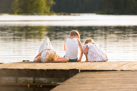 Traumhafter Sommer! Junge Frau sonnt sich auf einem Bootssteg und ihre beiden Kinder sitzen in der Sonne und beobachten etwas in Wasser. Sch&ouml;ne, entspannte Sommerstimmung, 