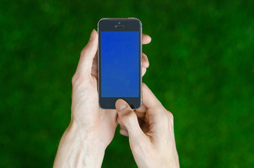 Human hand holding a modern mobile phone with a blue screen on a background of green grass first-person view