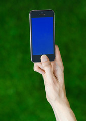 Human hand holding a modern mobile phone with a blue screen on a background of green grass first-person view