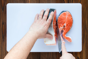 male hands cutting piece of salmon on a white cutting board on wooden background, top view.