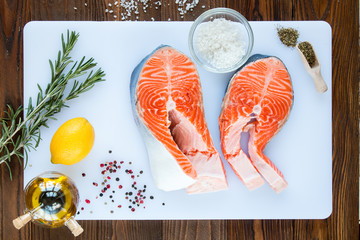 steaks of salmon with a sprigs of rosemary, lemon, pepper,  sea salt, olive oil and vinegar in glass jar, herbs in wooden scoop on a white cutting board on wooden background, top view.