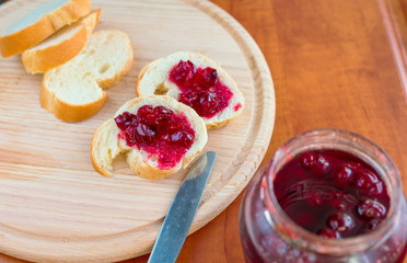 
Cherry jam in a glass jar with two slices of bread with jam and a knife on a wooden board