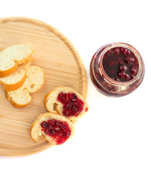 
Cherry jam in a glass jar with two slices of bread with jam and a knife on a wooden board on a white background .