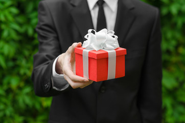 Gift and business theme: a man in a black suit holding a gift in a red box with a white ribbon on a background of green grass