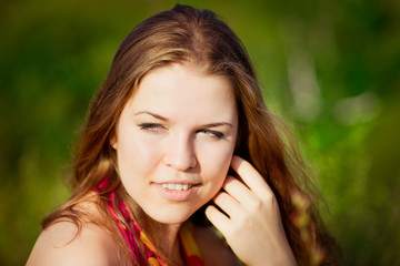 Fototapeta premium Close-up portrait of young woman with long red hair touching her face with her hand. Ginger girl sitting on grass in summer sunny field. Femininity concept