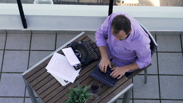 Young writer thinking, checking something on laptop while writing book on typewriter on terrace 
