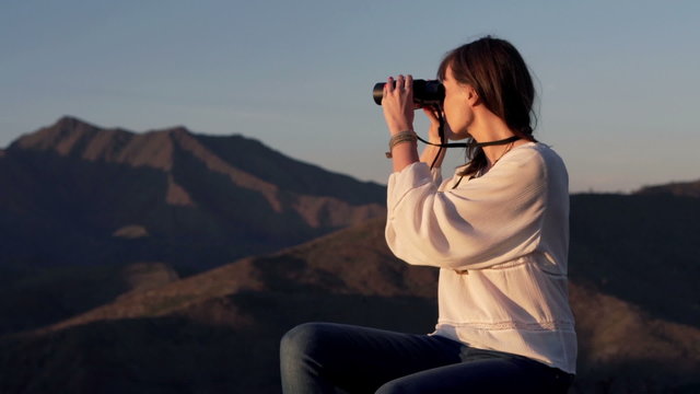 Young woman sitting on the trunk with binoculars and observing surroundings
