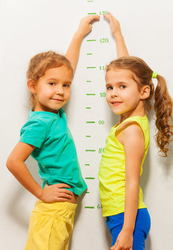 Two girls smile show height on wall scale at home