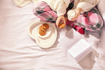 Fototapeta na wymiar Woman in pajamas reading a book and drinking milk with cookies on her bed