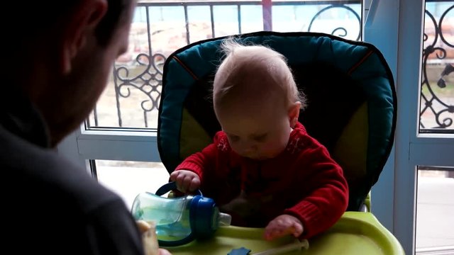 Young father feeding baby son in high chair in kitchen