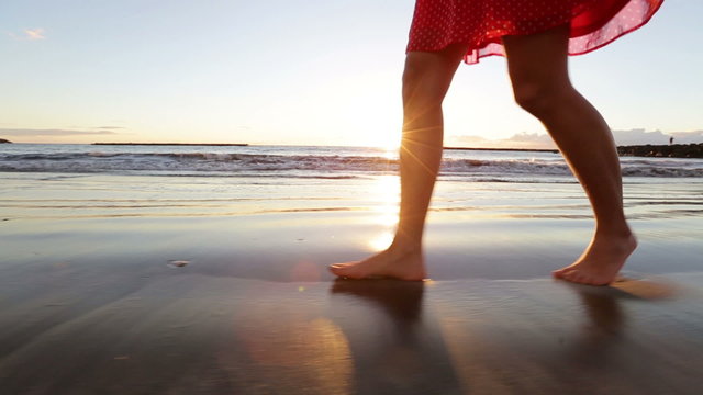 Woman feet and legs walking on beach at sunset. Closeup of beautiful girl in summer dress against the sun flare.