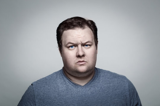 Close-up portrait of white Caucasian big fat stout man with blue eyes looking directly in camera in studio on light background, sceptical suspicious expression, emotion 