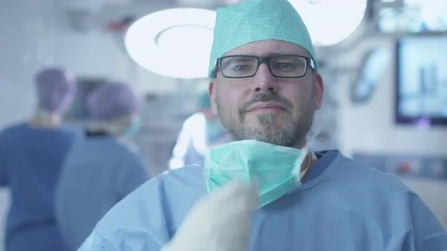 Portrait of Positive, Attractive and Smiling Doctor taking off Surgical Mask after successful Operation in Operating Room. Shot on RED Cinema Camera.