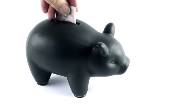 A young male inserts Euro, US Dollar and British Pounds into a black piggybank, then has enough of it and topples the piggybank.
