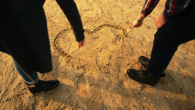 Young couple drawing heart on the sand by the wooden stick on the beach in cold weather in casual wear, sunset time, slow motion