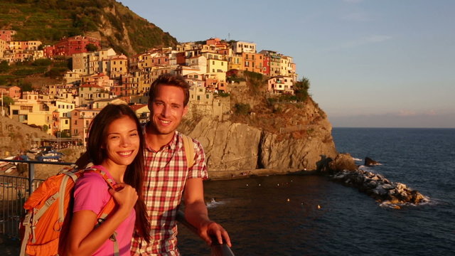 Hiking couple portrait looking at camera at sunset on holidays travel. Young backpacking Asian woman and Caucasian man enjoying ocean view on vacation in Manarola, Cinque Terre, Liguria, Italy